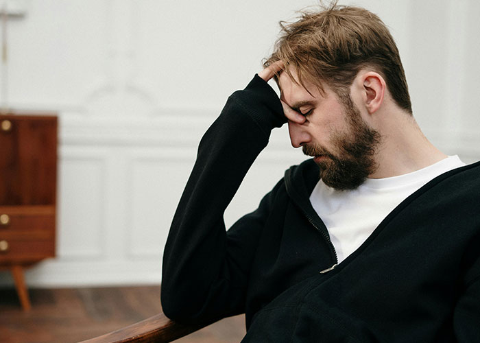 Man in a black hoodie sitting, appearing frustrated and thoughtful, hand on forehead in a home setting.