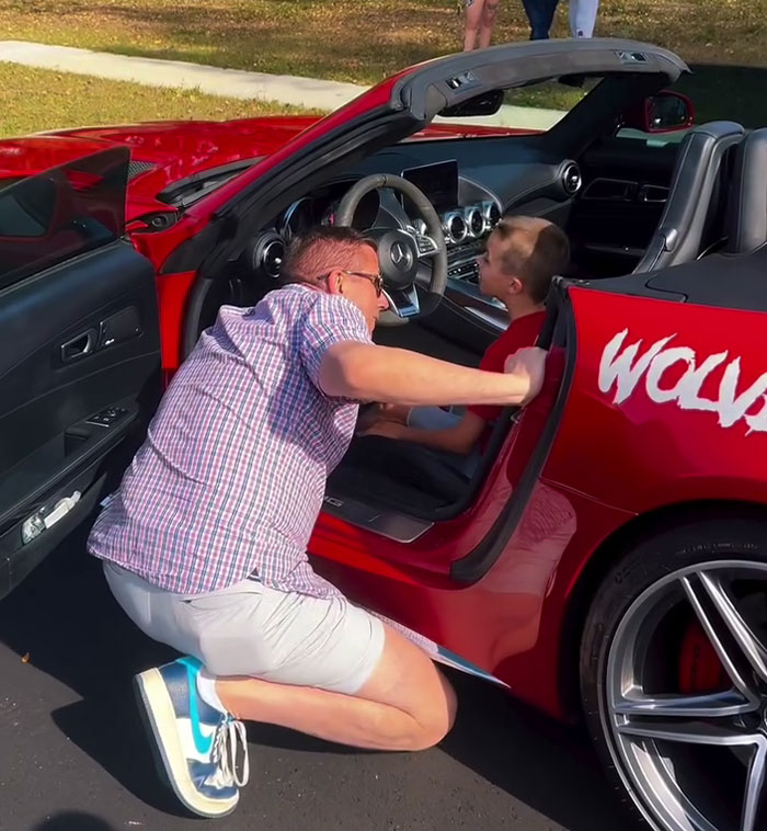 Boy sits in a red sports car as a person kneels beside, smiling during a birthday surprise from local car enthusiasts.