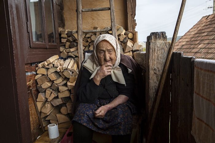 Elderly woman sitting on steps, surrounded by firewood, conveying the vulnerability of aging women.