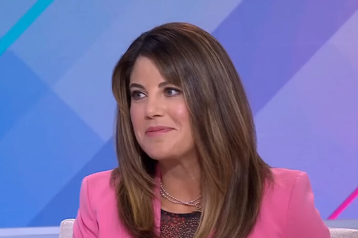 Celebrity in a pink blazer smiling during a TV interview, sitting against a colorful geometric background.
