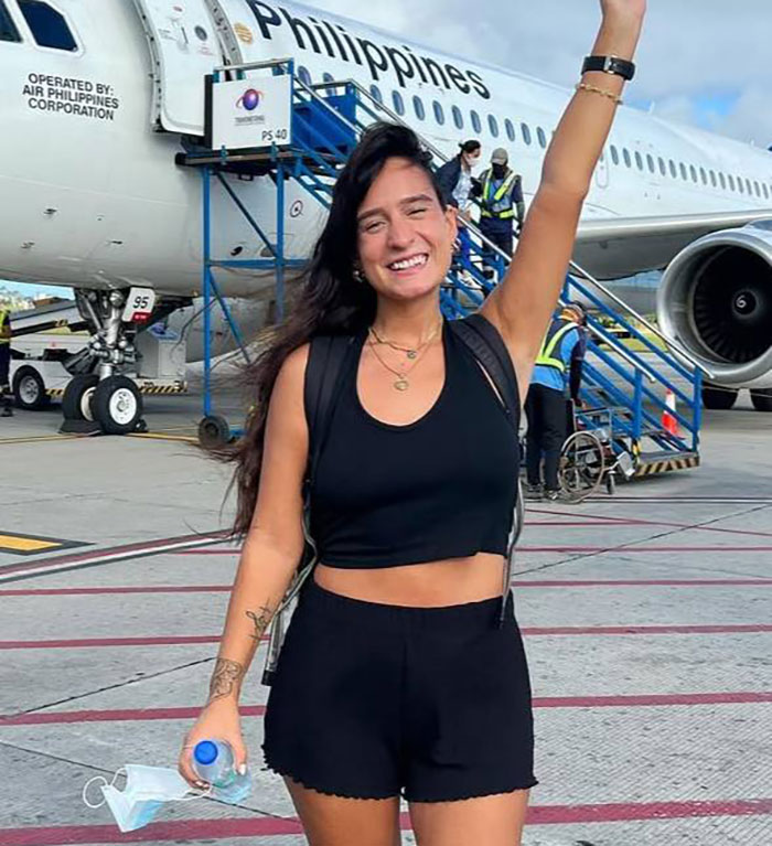 Smiling woman in black outfit stands in front of airplane, arm raised, before mysterious illness travel incident.
