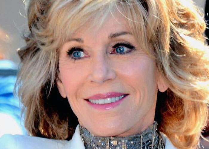 Smiling celebrity in white coat and glittery scarf at an outdoor event.