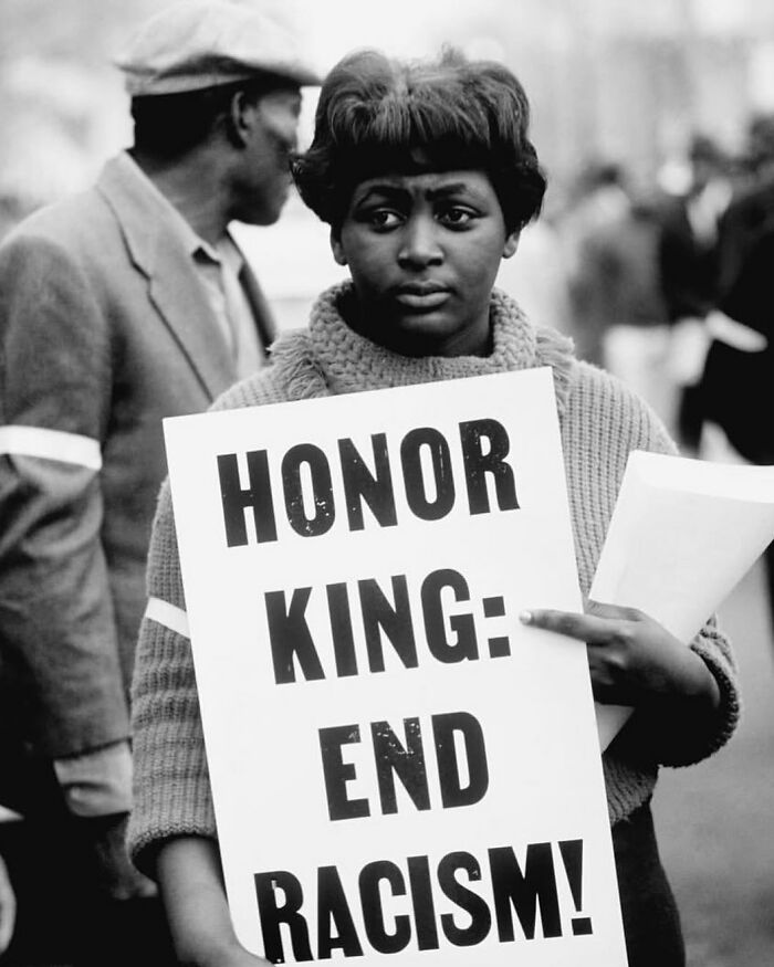Black and white street photo of a woman holding a sign protesting racism, featured on popular Instagram street photography page.