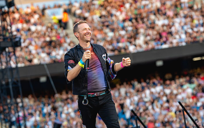 Celebrity smiling on stage during a live performance, wearing a colorful jacket, with a large audience in the background.