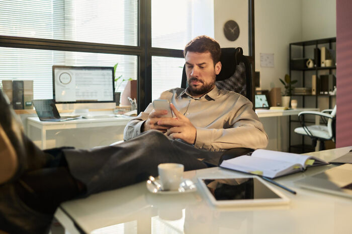Man in office chair with feet up, using smartphone, surrounded by work items. Unhinged work drama setting.