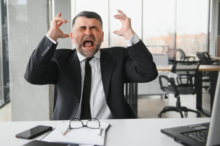 Frustrated man in suit yelling at desk, glasses and phone nearby, illustrating dramatic work scenario.