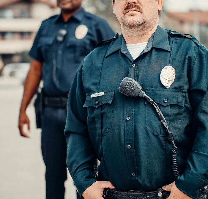 Two police officers in uniform standing outdoors, focus on one officer's badge and radio, representing authority and control.