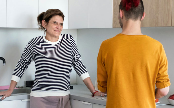 Woman in striped shirt in kitchen, looking surprised during a conversation about DNA results with family member in orange sweater.