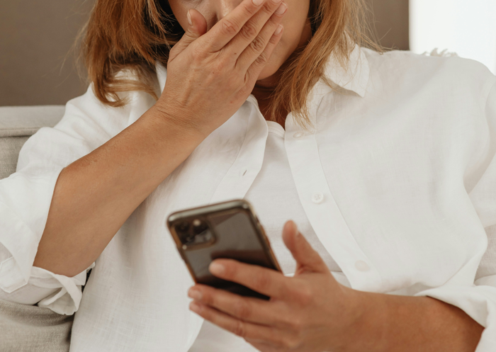 A woman in a white shirt looks shocked at her phone, covering her mouth.