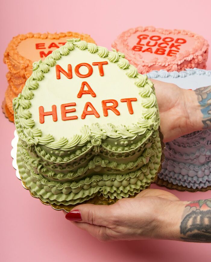 Decorated cakes with phrases like "NOT A HEART," held by tattooed hands, emphasizing vegan treats against a pink backdrop.