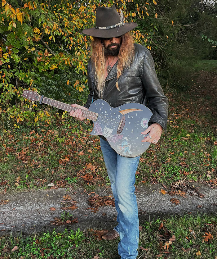 Man in a leather jacket and hat holding a decorated guitar, representing Billy Ray Cyrus' romance news.