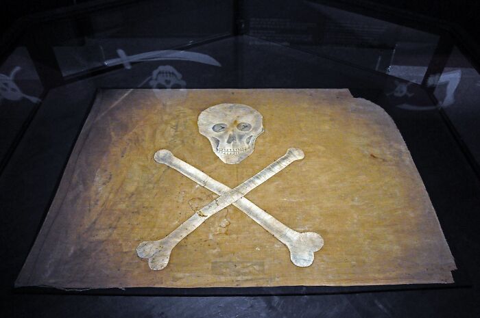 Historic artifact of a Jolly Roger pirate flag, featuring a skull and crossbones, displayed in a museum setting.
