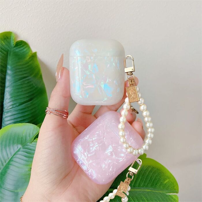 Hand holding iridescent and pink Amazon haul finds AirPods cases with pearl keychain accessories against green leaves.