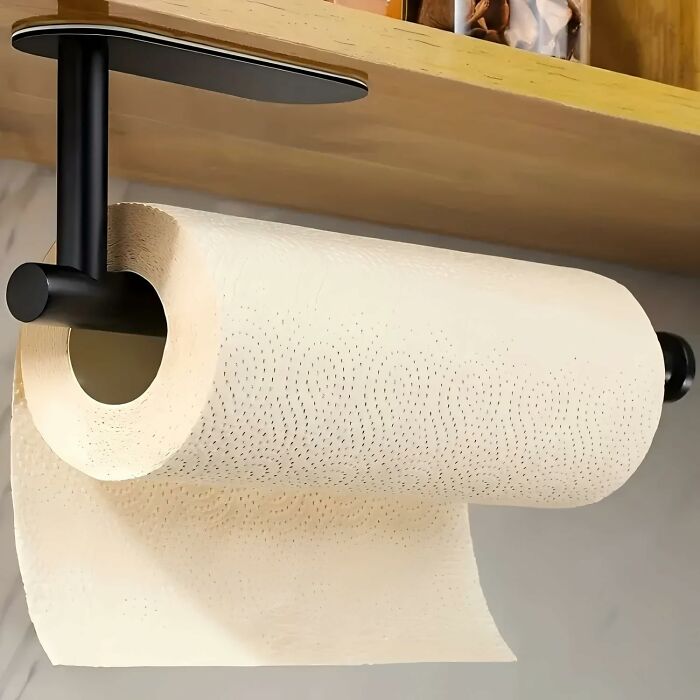 Black paper towel holder mounted under wooden kitchen shelf with a roll of paper towels hanging for easy access.