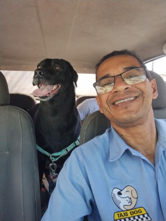 Man taking a selfie with a happy black dog in his pet taxi, wearing a light blue uniform with a "Taxi Dog" logo.