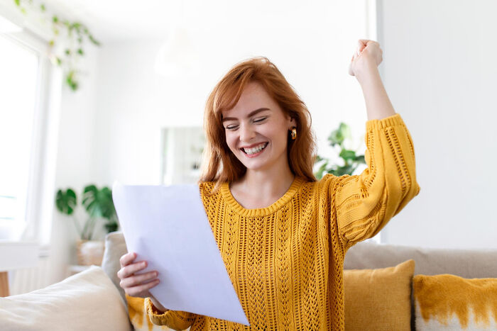 Happy woman celebrating with a raised fist while reading a paper, wearing a yellow sweater on a cozy couch.