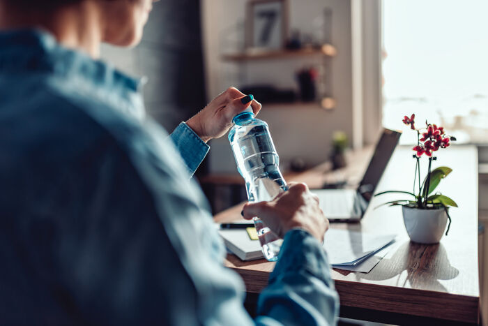 A person holding a water bottle in a home office, with a laptop and plant on the desk, creating a slightly creepy vibe.