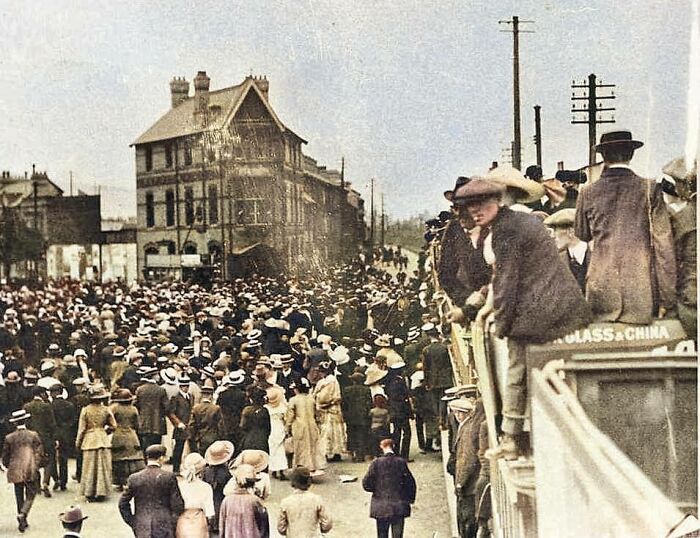 Crowd of people wearing early 20th-century clothing gathered on a European street, showing life in Europe 100 years ago.