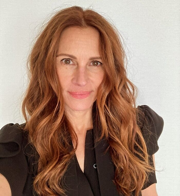 Celebrity posing with long wavy hair in a black top against a plain background.