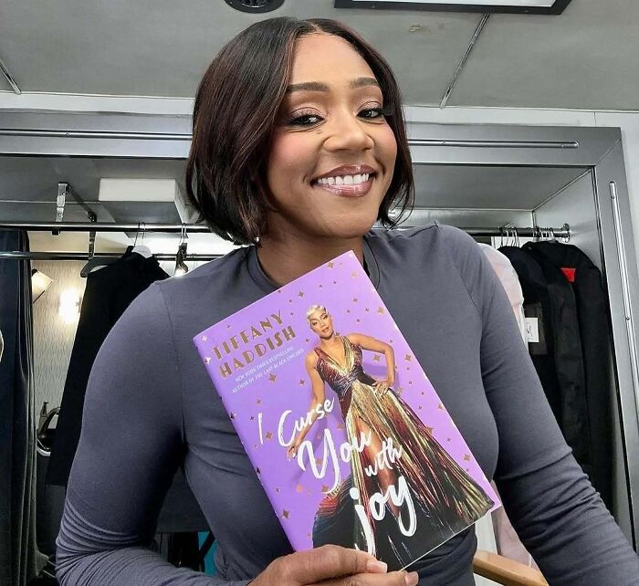 Smiling woman holding a book titled "I Curse You with Joy" in dressing room, showcasing petty celebrity moments.