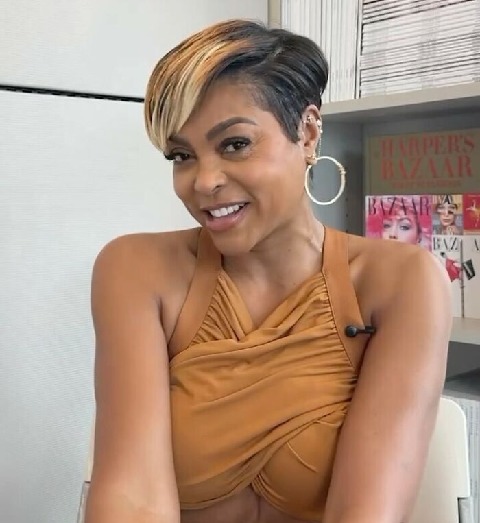 Celebrity smiling in a stylish dress, seated near a shelf with magazines in the background.