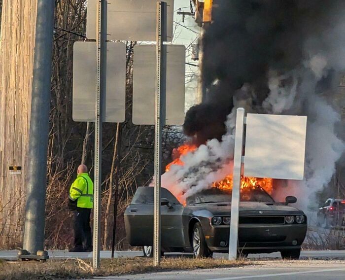 Car on fire at intersection, emitting heavy smoke while a person in a reflective jacket stands nearby, representing a horrible day.