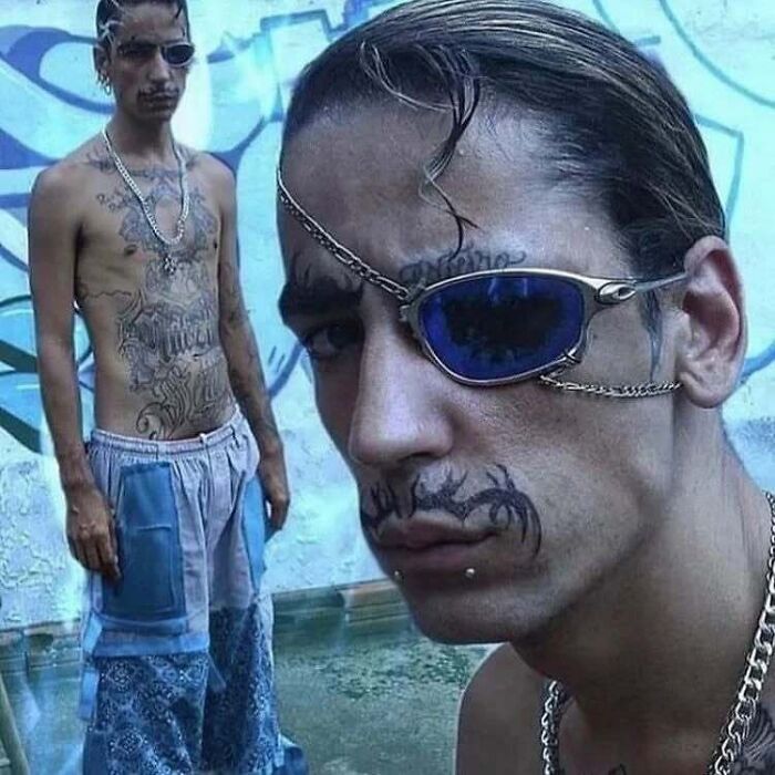 Two men with unique facial and body tattoos, one wearing sunglasses, standing against a graffiti wall.