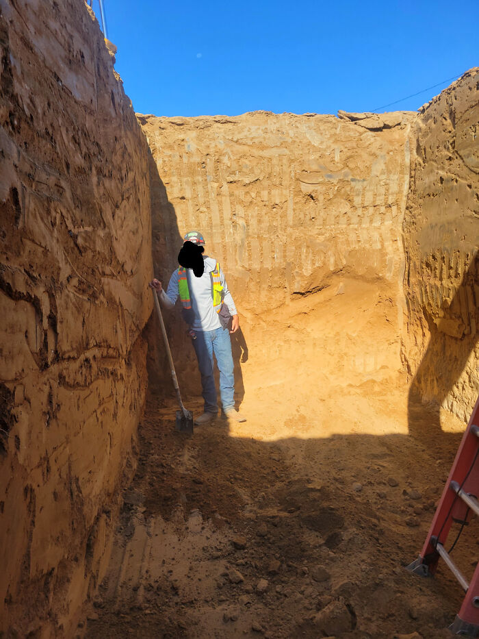 Worker standing in a deep trench, displaying a hazardous situation with steep walls and minimal safety equipment.