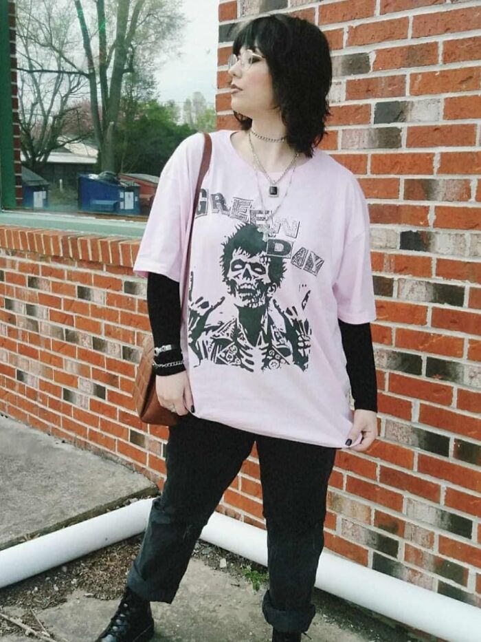 Person wearing Green Day shirt, black pants, and boots, standing by a brick wall. Festival survival kit fashion.