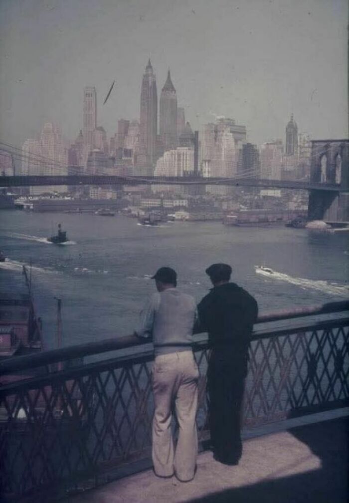 Two men leaning on a railing, overlooking a historical view of New York City with the Brooklyn Bridge in the background.