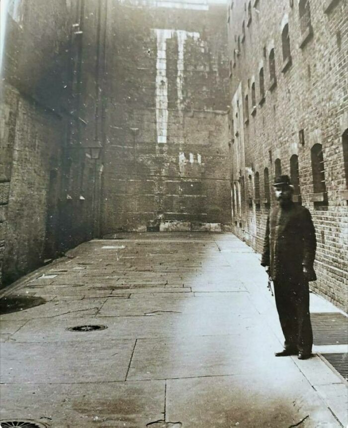 Old photo of a man standing in a narrow, brick-walled alley, evocative of historical urban architecture.