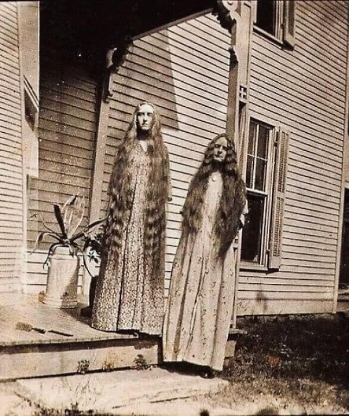 Two women with long hair stand on a porch in an old photo, showcasing historical fashion and style.