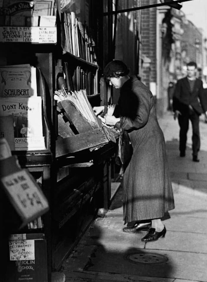 A woman examines sheet music outside a vintage store, capturing a moment in this historical photo.