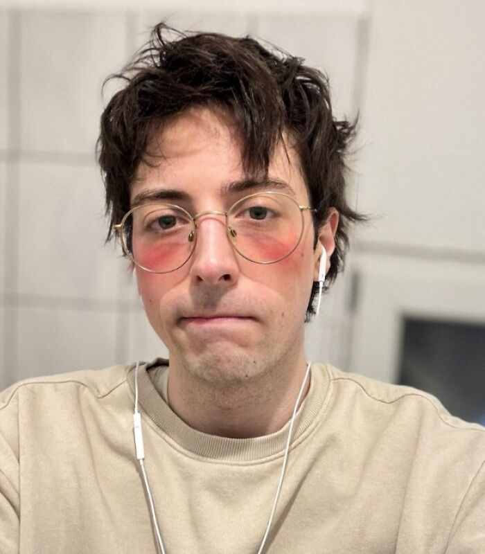 Joven con expresión de mal día usando gafas grandes y auriculares blancos en ambiente interior desenfocado.