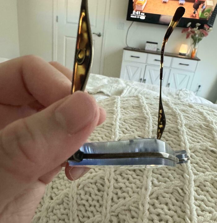 Bent glasses frame held in hand, background shows a TV on with a cozy room setting.