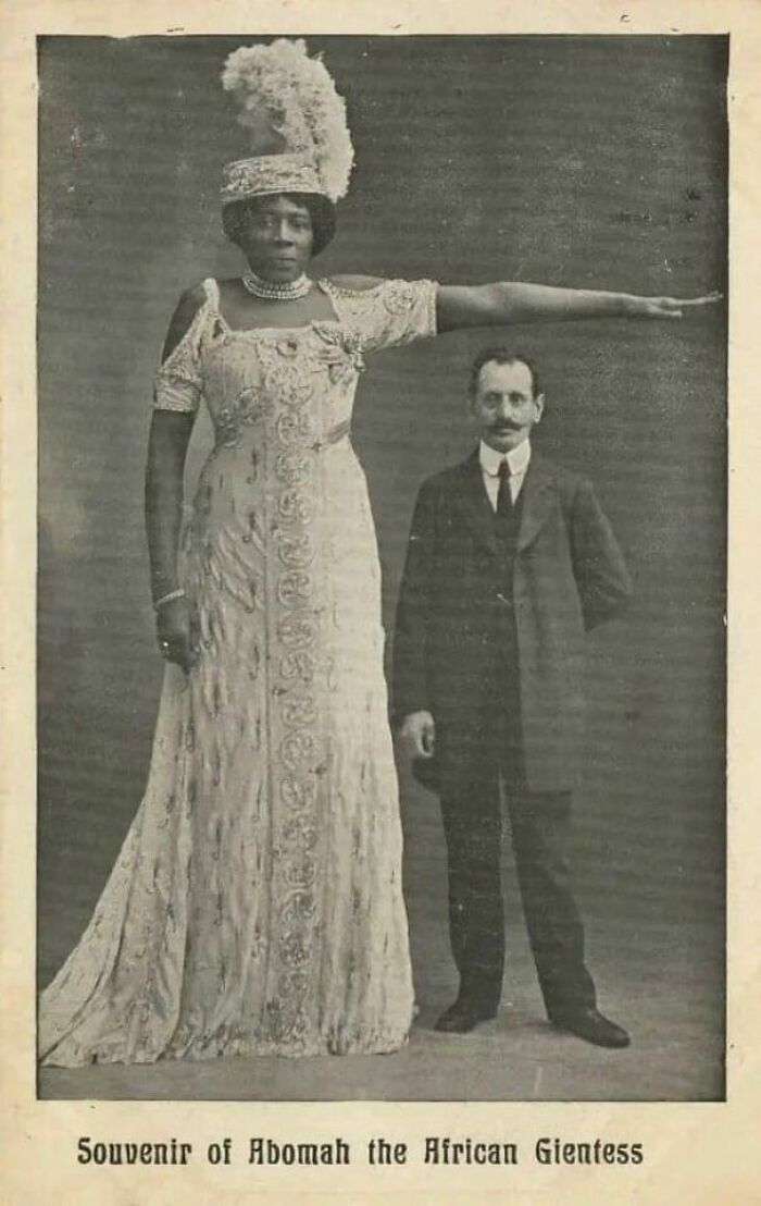 Tall woman in ornate dress with a feathered hat stands beside a man; historical photo for enthusiasts.