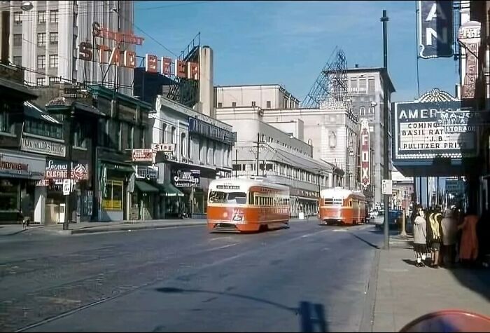 Vintage street scene with orange trams, historical buildings, and bustling sidewalks, capturing a moment in urban history.