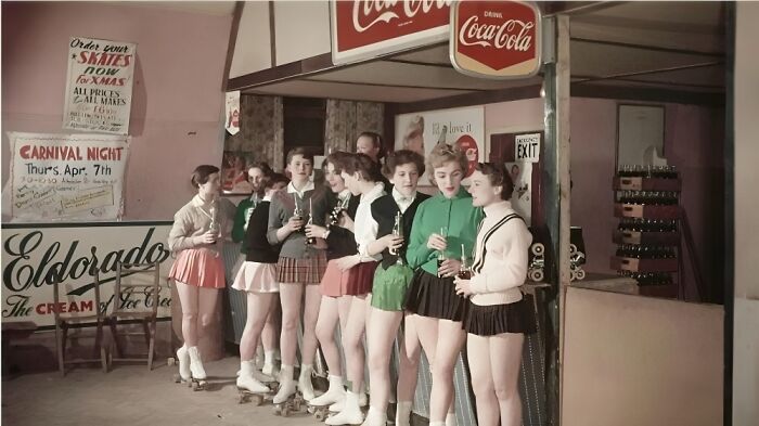 Women in vintage roller skate outfits holding drinks, standing under Coca-Cola signs at a historical skating event.
