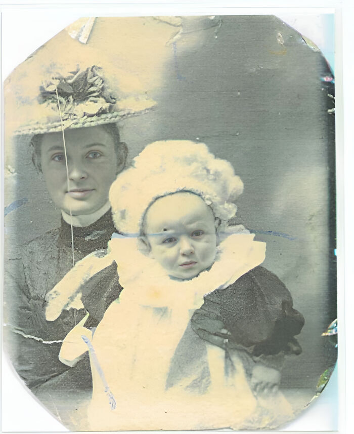 Vintage photo of a woman in a hat holding a baby in a white outfit, showcasing historical fashion styles.