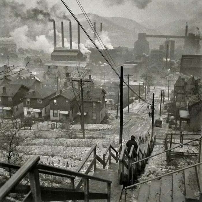 Old photo showing an industrial town with smoke rising from factories and a person walking down snowy steps.