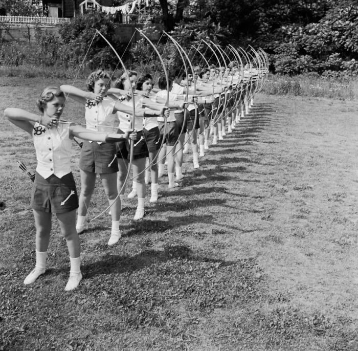 Women archers in a line preparing to shoot arrows, wearing matching uniforms, historical photo shared by enthusiasts online.