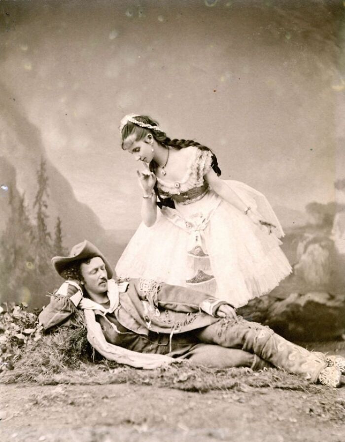A man in traditional attire reclines as a woman in an elegant dress stands over him in this fascinating old photo.