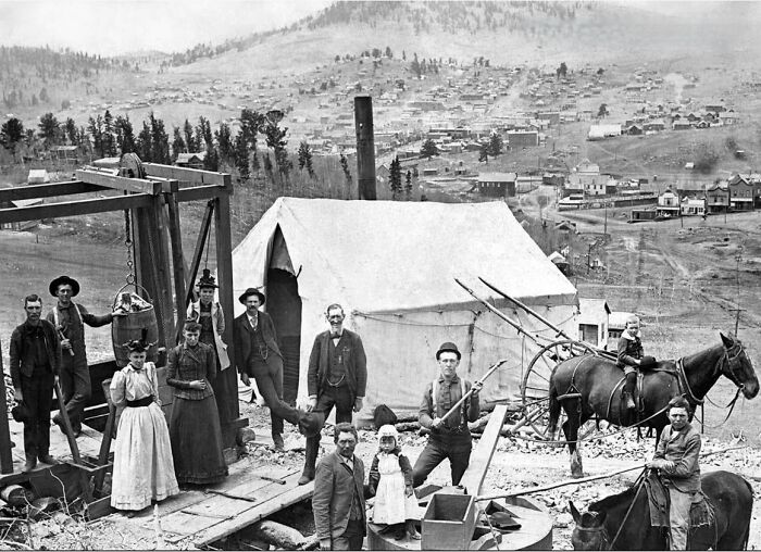 Historical photo of people and horses near a tented camp, with a panorama of a 19th-century town in the background.