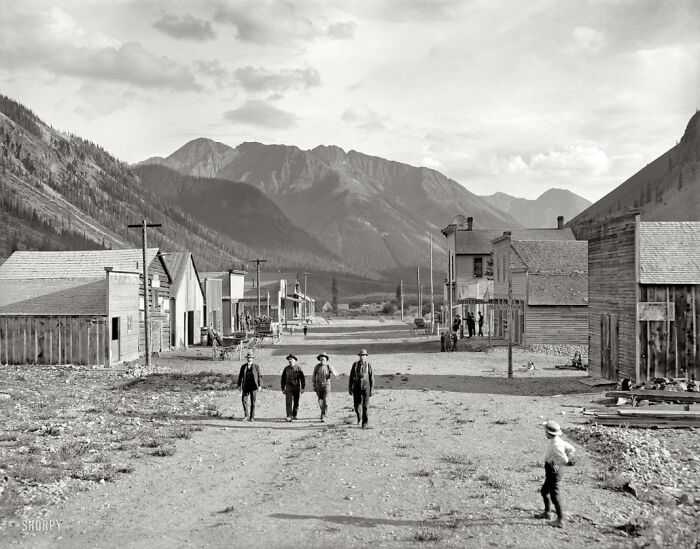 Historic town street with people walking, surrounded by mountains and wooden buildings.
