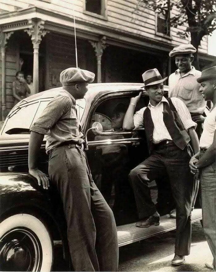 Four men in vintage attire lean on an old car, showcasing a moment of camaraderie.