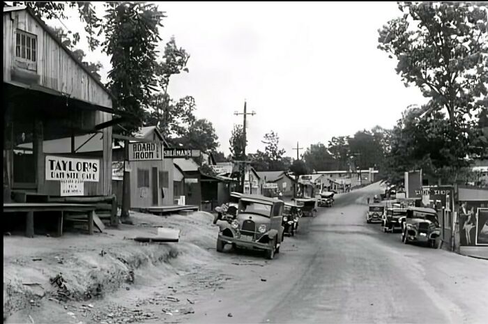 Historical photo of a vintage street scene with parked cars and old buildings lining the road.