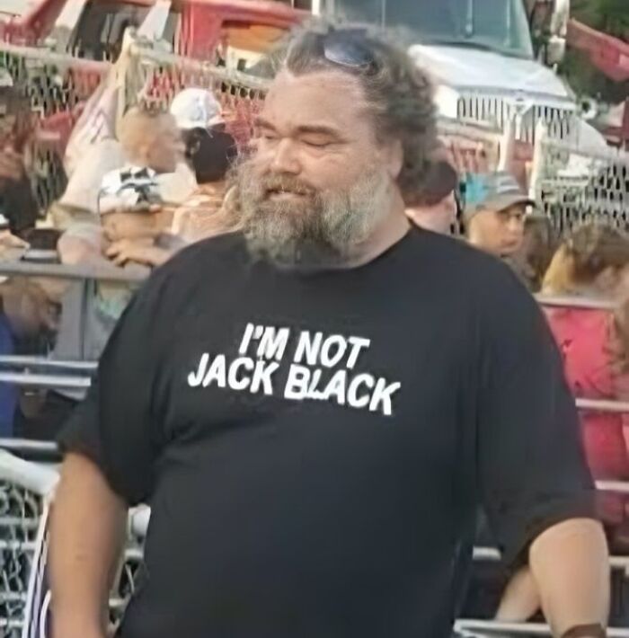 Hombre con camiseta negra divertida y ridícula con texto en inglés en evento al aire libre, entre público y vehículos.