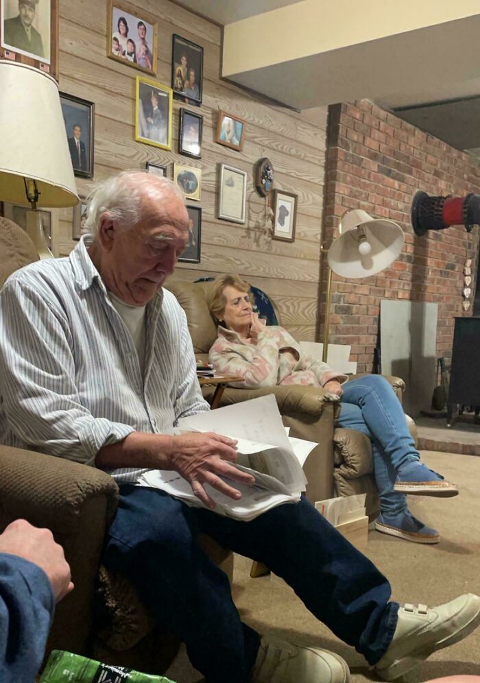 Elderly man reading papers, woman sleeping, humorous family room setting.