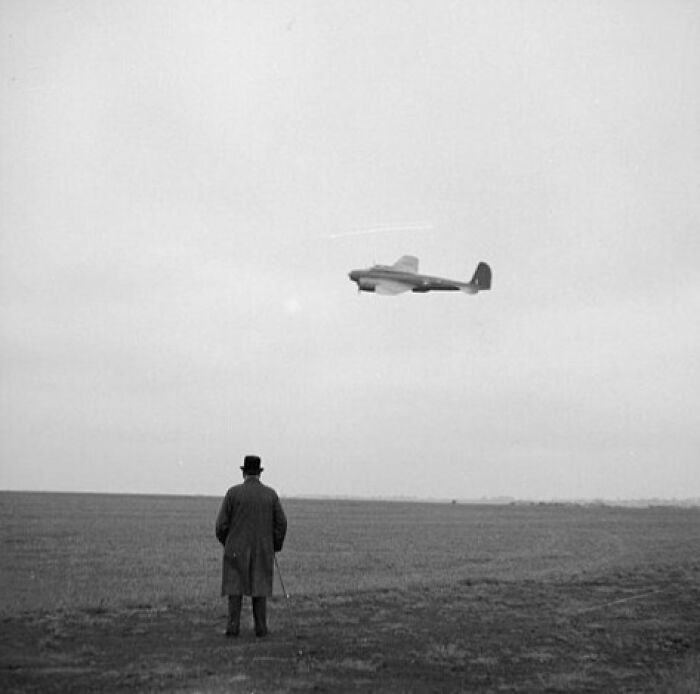 Man observing a WWII aircraft flying over an open field.