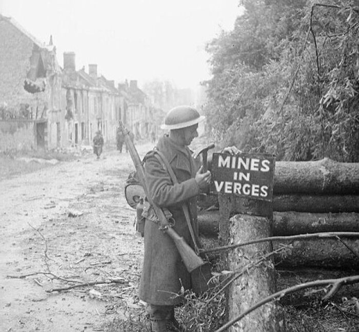 Soldier in WWII checking a sign reading "Mines in Verges" on a deserted street.